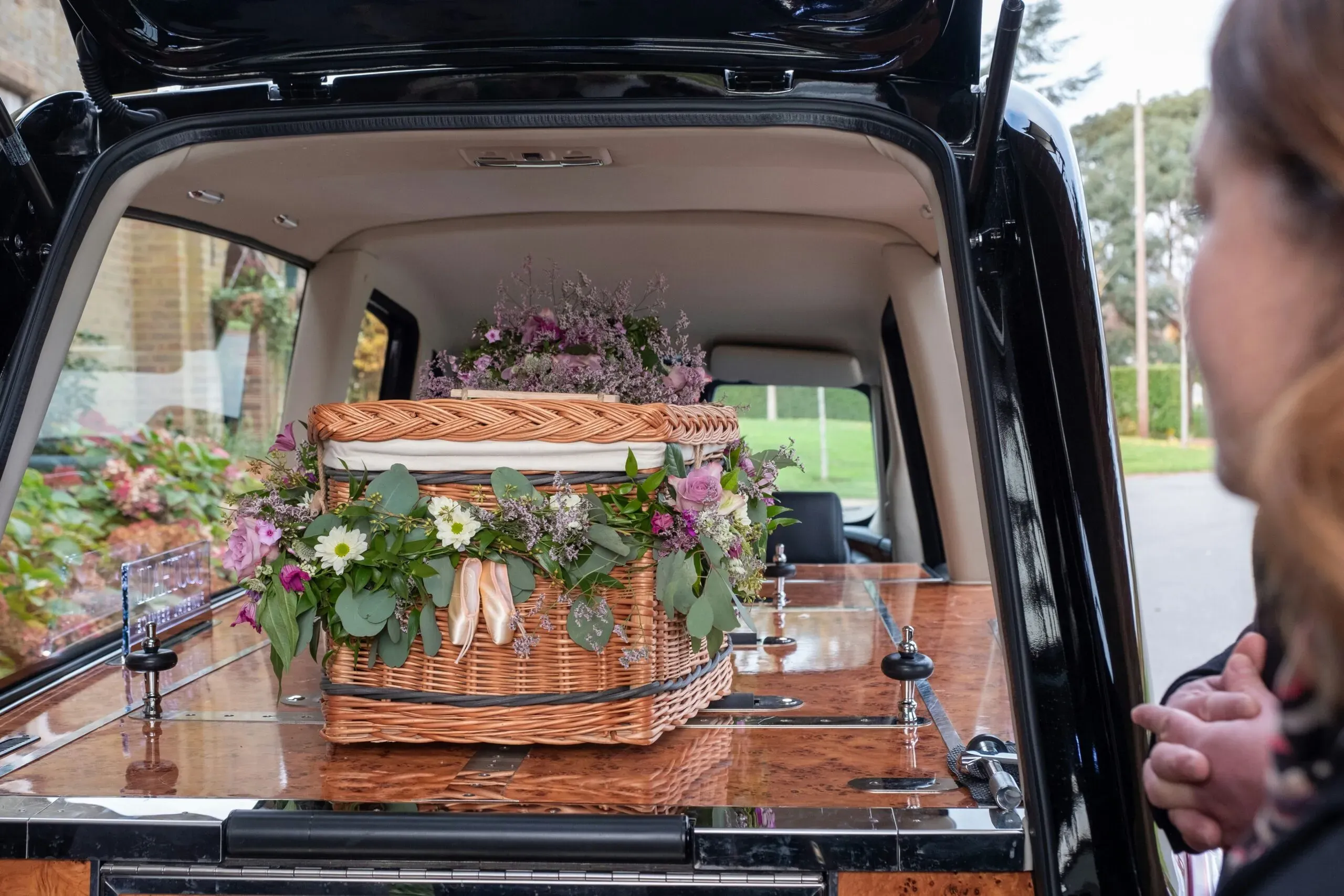 Cercueil en osier tressé fleuri à l'arrière d'un corbillard funéraire.